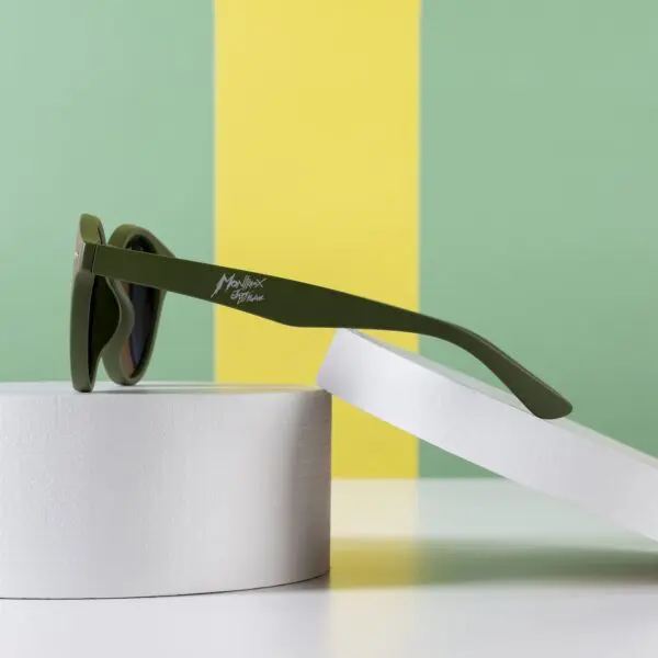 Fly Bruxelles Khaki Glasses displayed on a white circular stand against a green and yellow color-blocked background. The side-on angle highlights the khaki frame and the text on the arm.