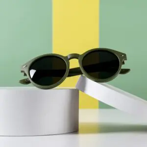 The Fly Bruxelles Khaki Glasses, featuring black lenses and round green frames, are displayed on two white cylindrical platforms against a green and yellow striped background.