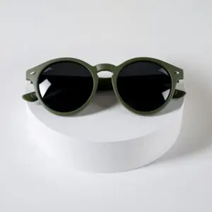 A pair of Fly Bruxelles Khaki Glasses with round, olive green frames and dark lenses, showcased on a white cylindrical platform against a plain white background.