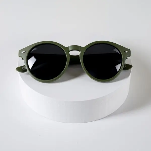 A pair of Fly Bruxelles Khaki Glasses with round, olive green frames and dark lenses, showcased on a white cylindrical platform against a plain white background.