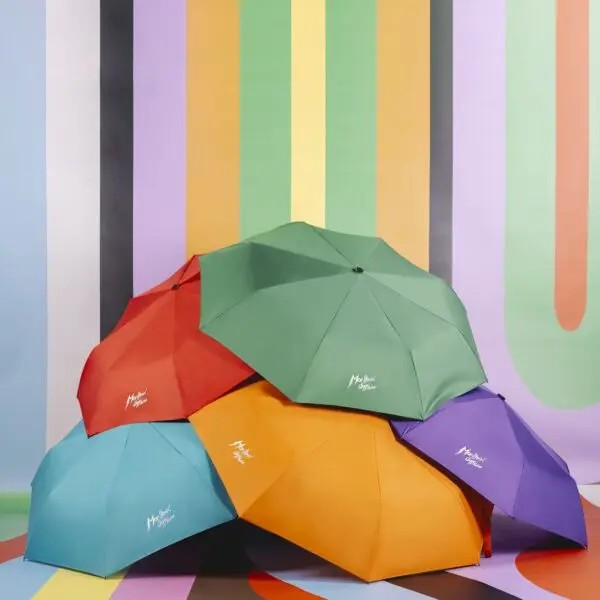 Cinq parapluies Parapluie colorés - rouge, vert, orange, violet et bleu - sont empilés devant une toile de fond rayée multicolore et vibrante, chacun portant la marque blanche du Montreux Jazz.