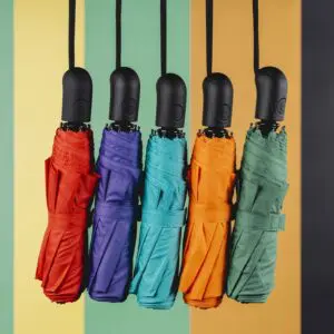 Cinq parapluies Parapluie rouges, violets, bleus, orange et verts sont suspendus sur un fond rayé jaune, vert et orange. Leurs poignées noires sont régulièrement espacées, reflétant l'esprit ludique de la collection Parapluie.