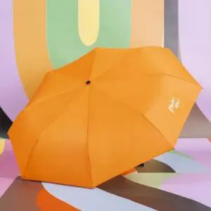 Un parapluie ouvert en orange vif avec un texte blanc sur un panneau se trouve sur un fond abstrait coloré avec des formes courbes en vert pastel, violet, orange et gris.