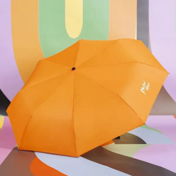 Un parapluie ouvert en orange vif avec un texte blanc sur un panneau se trouve sur un fond abstrait coloré avec des formes courbes en vert pastel, violet, orange et gris.