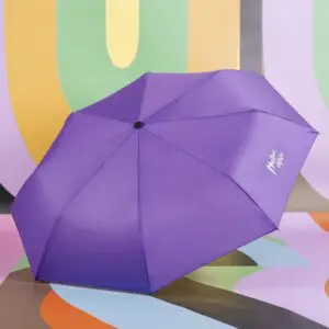 Un parapluie Parapluie violet portant l'inscription "Kashmir Diaries" en blanc sur un panneau est ouvert et présenté sur une surface et un fond colorés à motifs abstraits inspirés du Montreux Jazz.