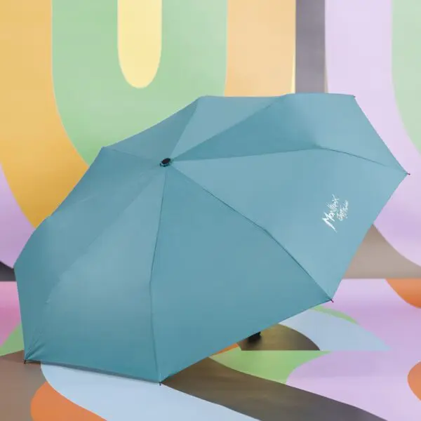 Un parapluie Parapluie sarcelle avec "Michael Soffe" en blanc est ouvert sur un fond abstrait inspiré de Parapluie Couleurs Montreux Jazz, avec des formes tourbillonnantes vertes, jaunes, orange et violettes.