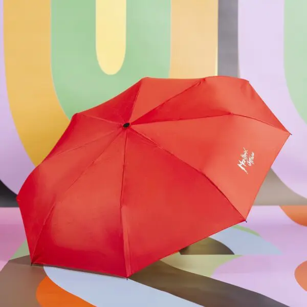Un parapluie Parapluie rouge vif avec un texte blanc sur un panneau se tient ouvert sur une toile de fond abstraite et vivante présentant des formes courbes en vert, jaune, orange, violet et noir.