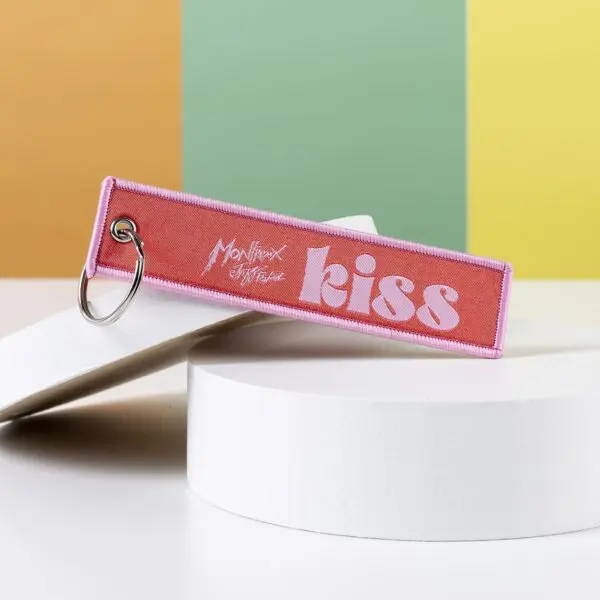 Porte-clés Kiss en tissu rose avec "Montreux Jitterbug" et "kiss" en texte blanc, présenté sur une plateforme blanche avec des rayures jaune pastel, vertes et orange - idéal comme accessoire de Montreux Jazz.