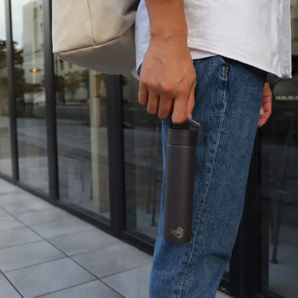 Une personne vêtue d'un jean bleu et d'une chemise blanche se tient à l'extérieur, près de grandes baies vitrées. Elle porte un sac fourre-tout beige et tient une Mini Bouteille Isotherme de 180 ml.