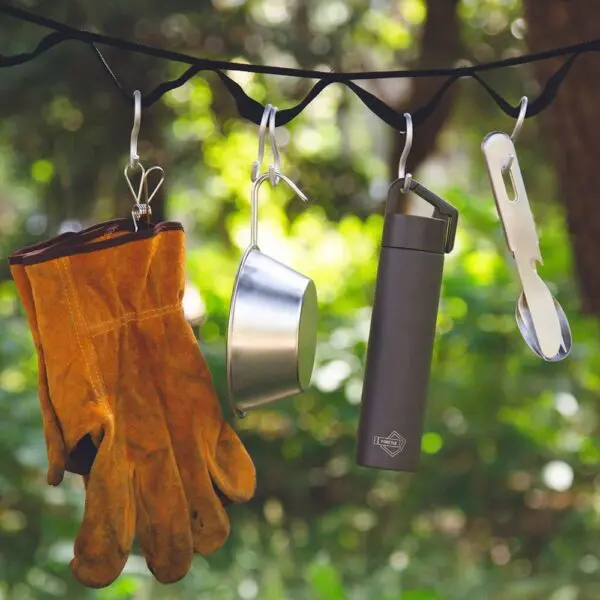 Une paire de gants de travail bruns, une tasse en métal, une Mini Bouteille Isotherme 180ml et un ustensile blanc sont suspendus à des crochets sur une sangle noire en extérieur, avec un feuillage vert flou en arrière-plan.