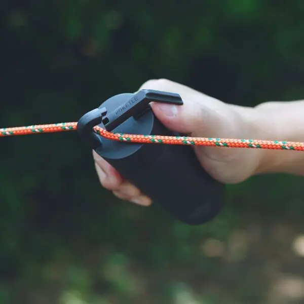 Une main attache une corde orange et verte à un dispositif cylindrique noir muni d'un crochet, tandis qu'une Mini Bouteille Isotherme 180ml repose à proximité sur un fond extérieur flou.
