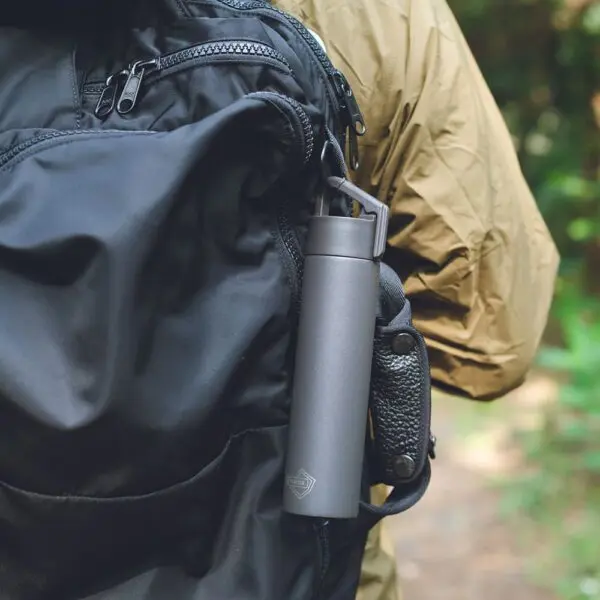 Une Mini Bouteille Isotherme de 180 ml de couleur grise est accrochée à un sac à dos noir porté par une personne vêtue d'une veste vert olive, sur fond de forêt floue.