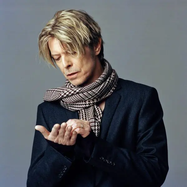 Une personne aux cheveux blonds ébouriffés, vêtue d'un blazer noir et d'une écharpe à carreaux, regarde pensivement sa paume ouverte, évoquant le style introspectif du Coffret David Bowie 6 : I Can't Give Everything Away (2002-2016).