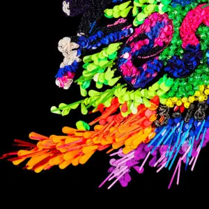 Une œuvre d'art abstraite vibrante inspirée par Affiche Kévin Germanier, 2026, composée d'objets en plastique colorés, de perles et de paillettes sur fond noir avec des motifs néon tourbillonnants en vert, rose, orange, jaune, violet et bleu.