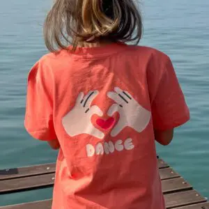 Un enfant se tient sur un quai en bois face à l'eau, portant le "T-Shirt Dance, Enfant" en corail, avec deux mains illustrées formant un cœur et "DANCE" imprimé au dos.