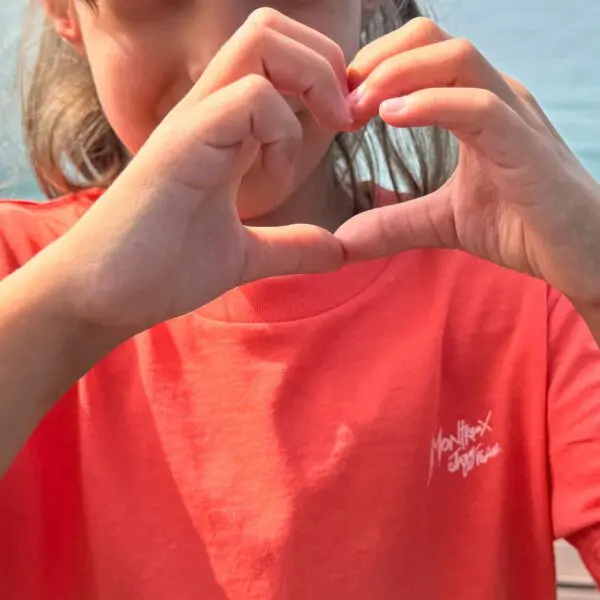 Un enfant portant le t-shirt Dance, Enfant in coral forme un cœur avec ses mains. L'arrière-plan flou représente l'eau et la lumière du soleil, tandis que le visage de l'enfant est partiellement hors du cadre.