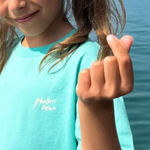Un enfant vêtu d'un T-Shirt Love, Enfant bleu clair se tient au bord de l'eau et fait le geste du cœur avec sa main droite.