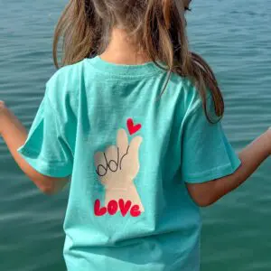 Un enfant aux cheveux longs portant le T-Shirt Love, Enfant se tient au bord de l'eau. Le tee-shirt montre une main avec le signe "je t'aime", un cœur rouge au-dessus, et "LOVE" écrit en dessous.