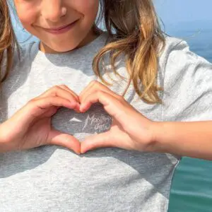 Une jeune fille blonde avec des nattes forme un cœur avec ses mains sur son T-Shirt Music, Enfant, souriant doucement en plein air dans un cadre lumineux près de l'eau.