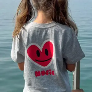 Une jeune fille avec des nattes se tient sur un quai et regarde l'eau calme. Elle porte le t-shirt Music, Enfant, un t-shirt gris avec un cœur rouge, un sourire noir et le mot "music" imprimé au dos.