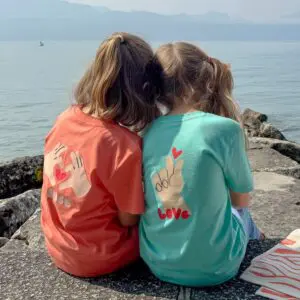 Deux jeunes filles sont assises côte à côte sur des rochers au bord d'un lac, face à l'eau. Elles portent des chemises colorées avec des signes de la main et les mots "Peace" et "Love" imprimés dans le dos. Des montagnes sont visibles dans l'arrière-plan brumeux.